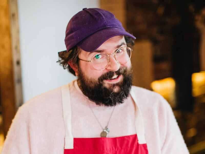 A man wearing a pink tshirt and purple baseball cap is smiling at the camera for a photo